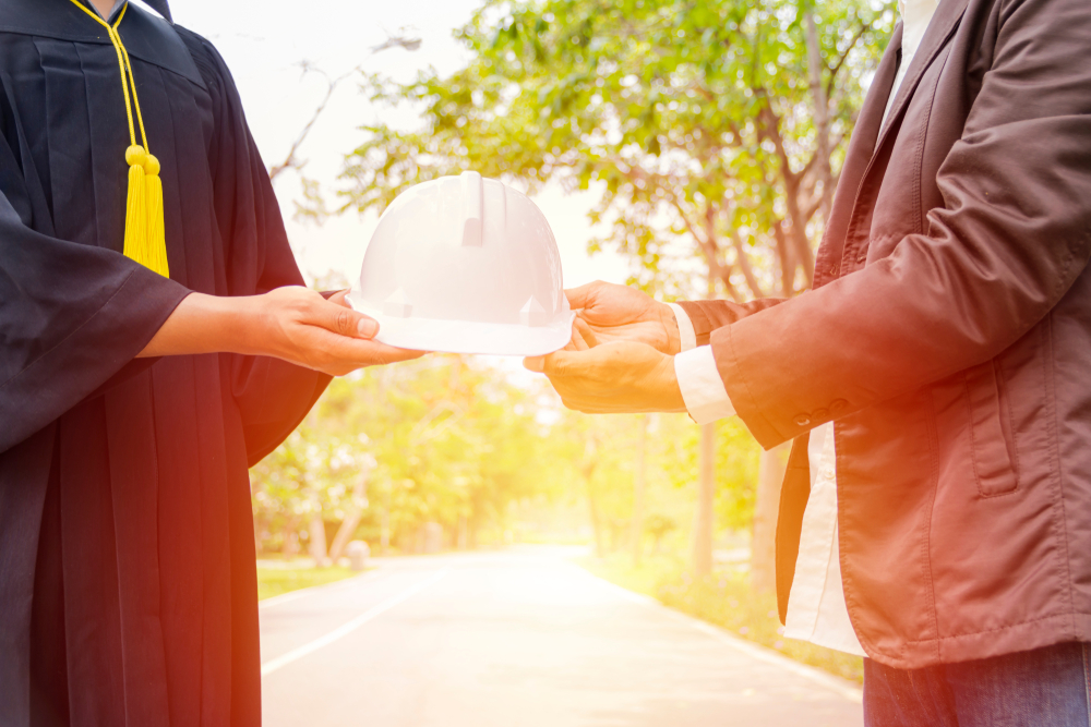 two men holding a hard hat between them, with one wearing a graduation gown and the other wearing a suit