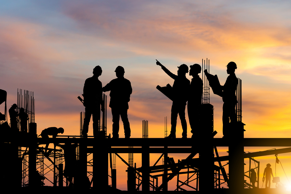 silhouettes of construction workers standing on top of structural beams with sunset in background