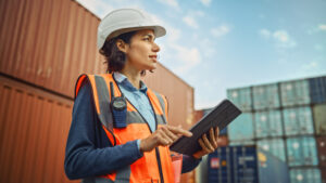 Female engineer smiles in hard hat and high visibility vest, working on tablet.
