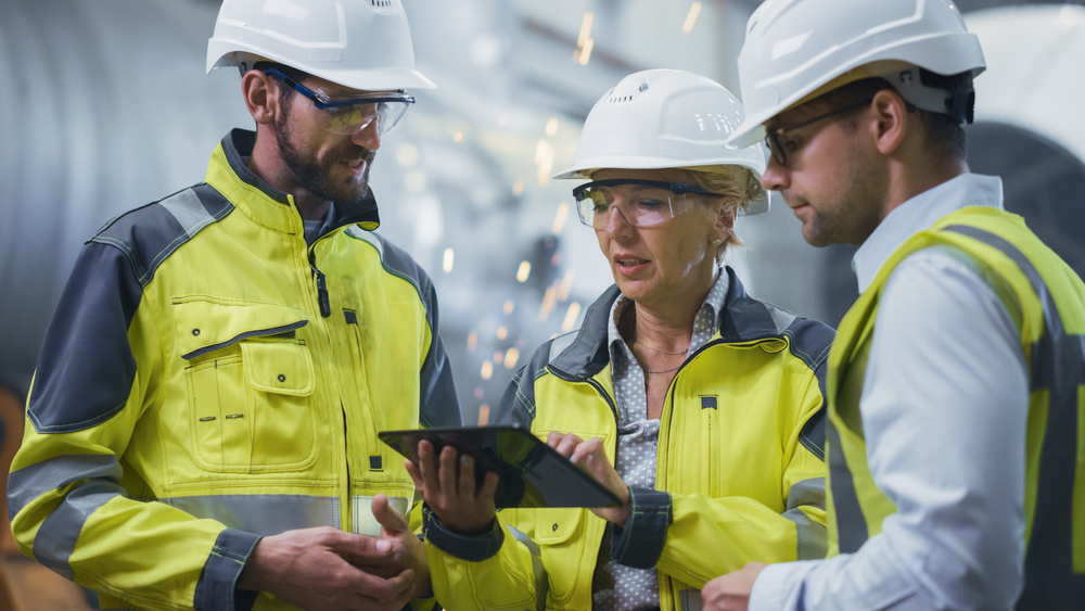 Three engineers in safety gear have a discussion in a manufacturing facility.