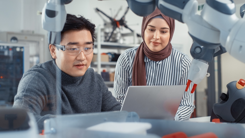 In Technology Research Facility: Young Female Specialist Talks With Asian Engineer. They Have A Discussion and Point at Laptop Screen. Professionals Work on Autonomous Factory Optimisation.